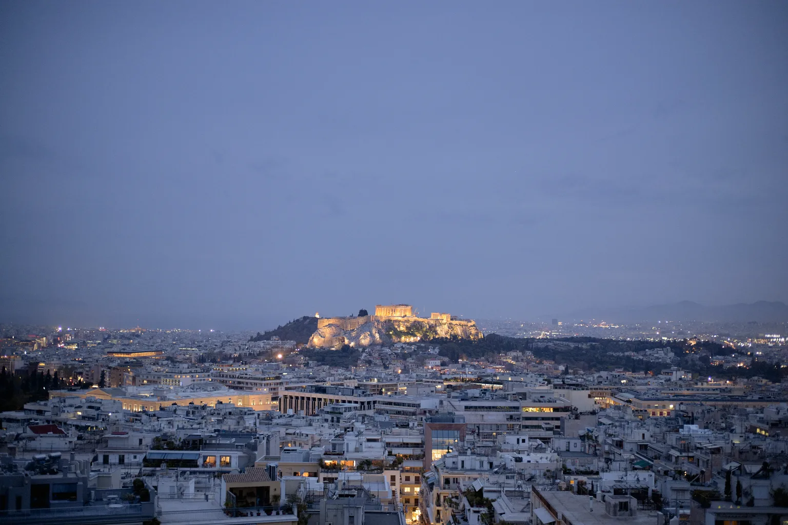 Αεροφωτογραφία της Αθήνας στο σούρουπο, με τον φωτισμένο Παρθενώνα και την Ακρόπολη πάνω από την πόλη.
